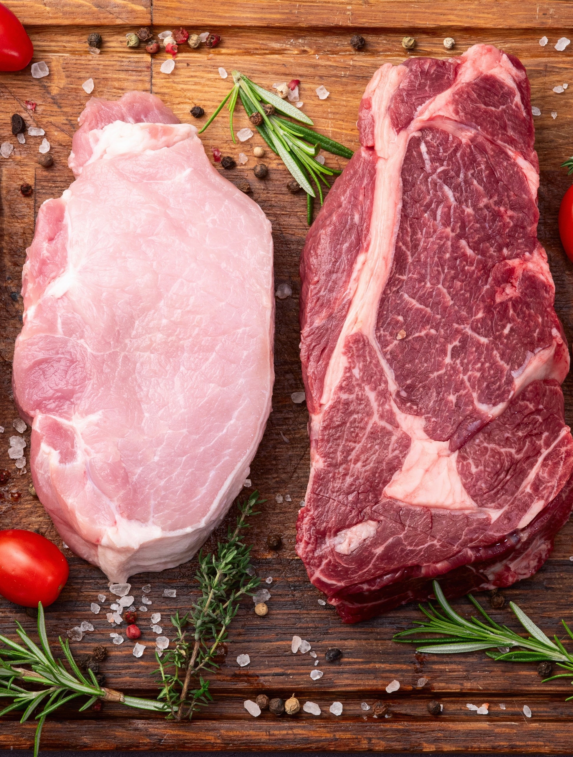 Two raw cuts of pork and beef on a wooden board, garnished with rosemary and cherry tomatoes.